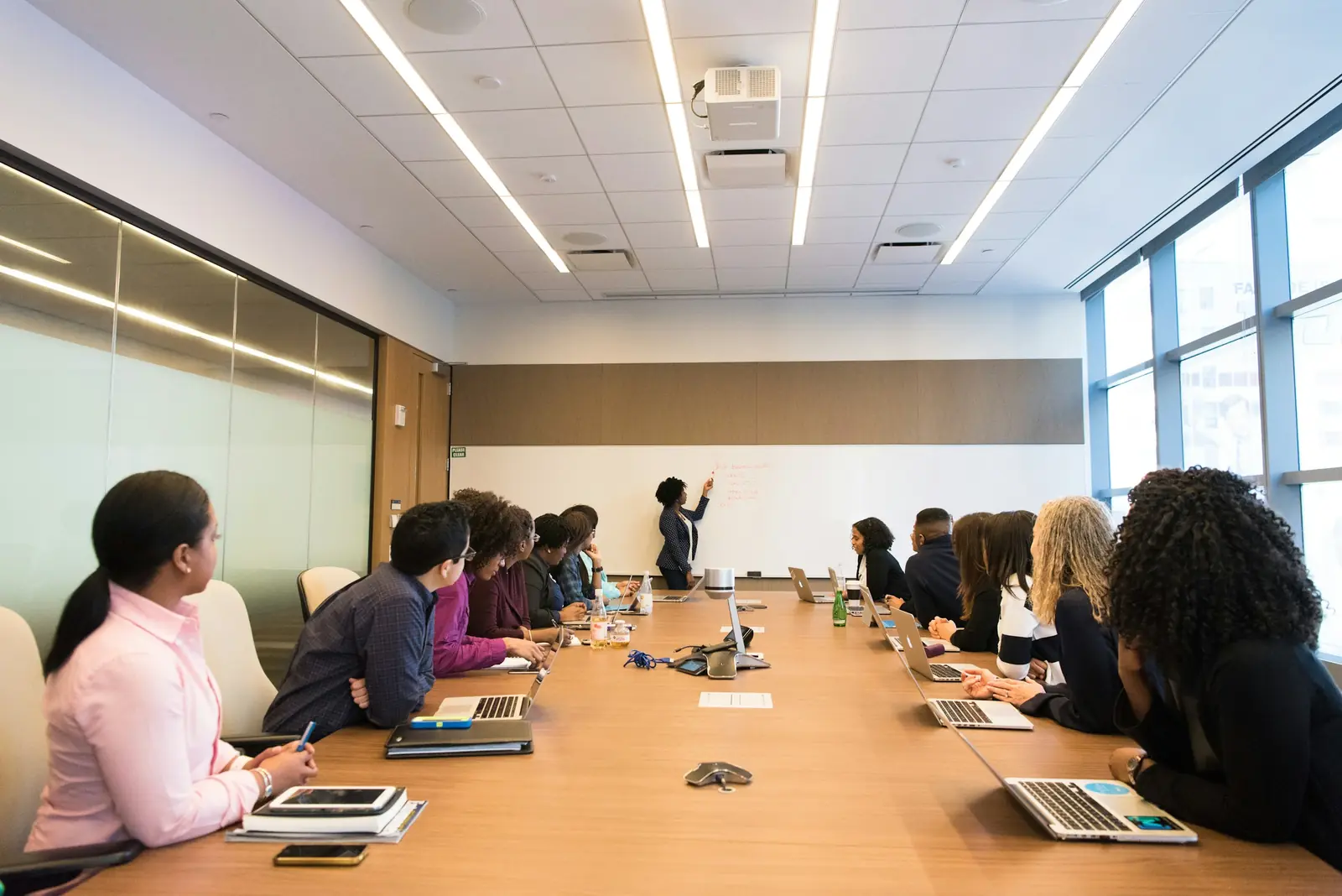 Equipa diversificada reunida à volta de uma mesa de conferência, enquanto uma mulher apresenta no quadro branco num escritório moderno